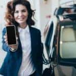 A woman is holding her phone out while standing in front of her car she was able to get from no credit car loans in Columbus.