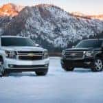A 2015 Chevy Tahoe is parked next to a 2015 Suburban with mountains in the background.