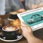 A person is searching on their laptop for a new car in a coffee shop after researching 'can I trust buying a car online?'