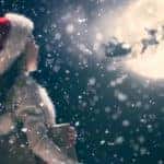 A small child is holding a book while watching Santa's slay cross over the moon at night.
