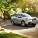 A silver 2017 Subaru Forester is driving on a sunny, tree-lined street.