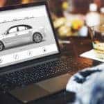 A man is having a drink while looking for the best time to buy a car online in Columbus, OH, on his laptop.
