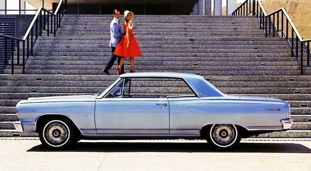 A 1960s couple is walking up stairs behind a light blue 1964 Chevy Malibu.