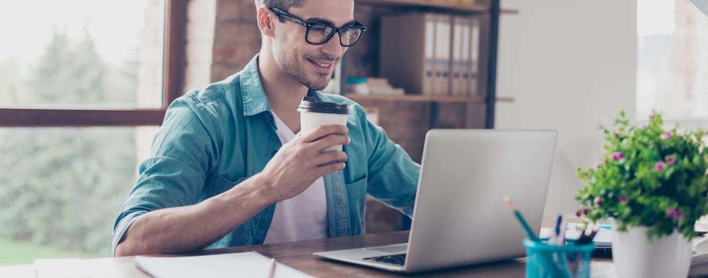 A man is smiling while holding a coffee and using his laptop.