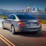 A silver 2019 Honda Accord is shown from the rear driving next to a river and city at night.