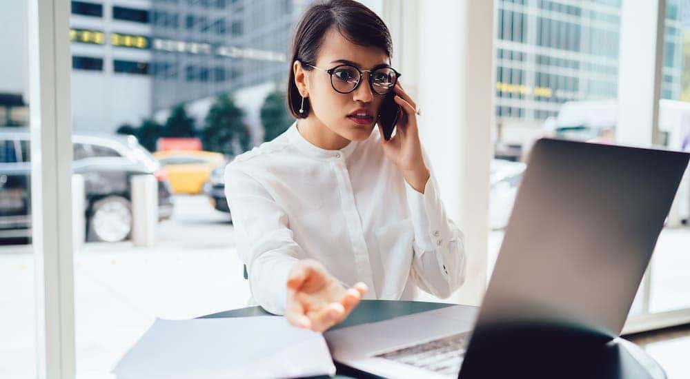 A frustrated business woman is inquiring about bad credit car loans while on a phone and laptop.