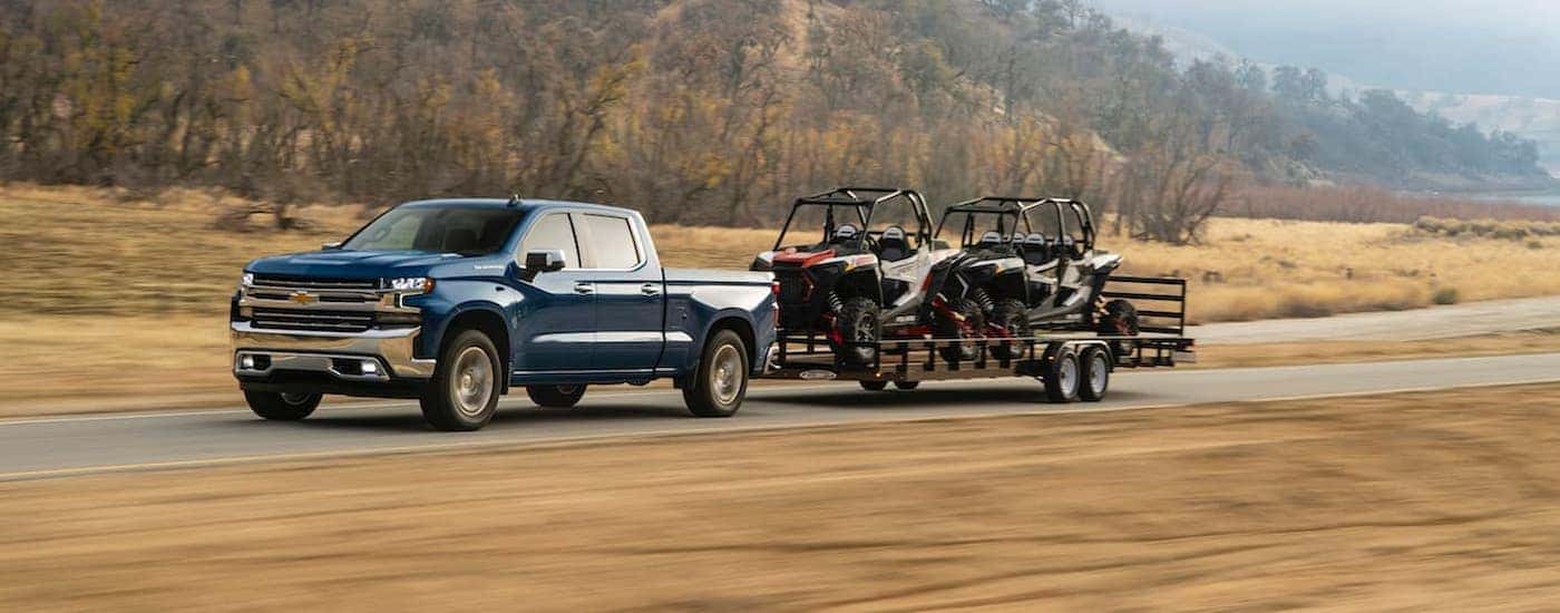 A blue 2020 Chevy Silverado 1500 is shown towing two UTVs on a small trailer.