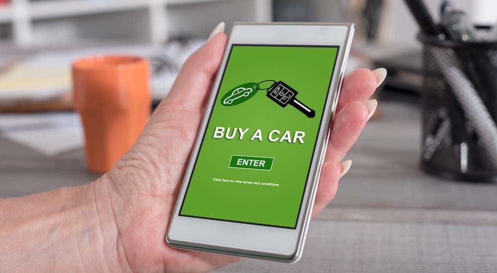 A woman's hand is holding a cell phone and reads "buy a car" as she shops at an online car dealer.