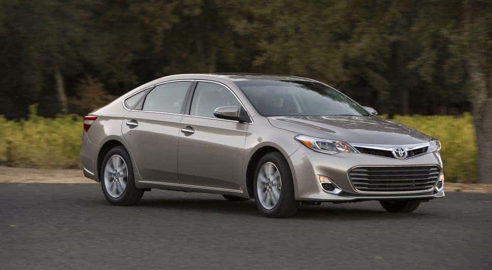 A popular used car in Indianapolis, a gray 2013 Toyota Avalon, is driving on a tree-lined road.