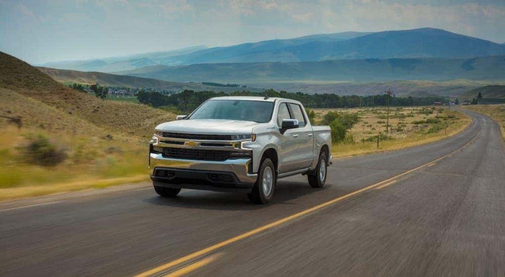 A popular used truck for sale in Kentucky, a silver 2020 Chevy Silverado 1500 LT, is driving on an empty highway in front of distant mountains.