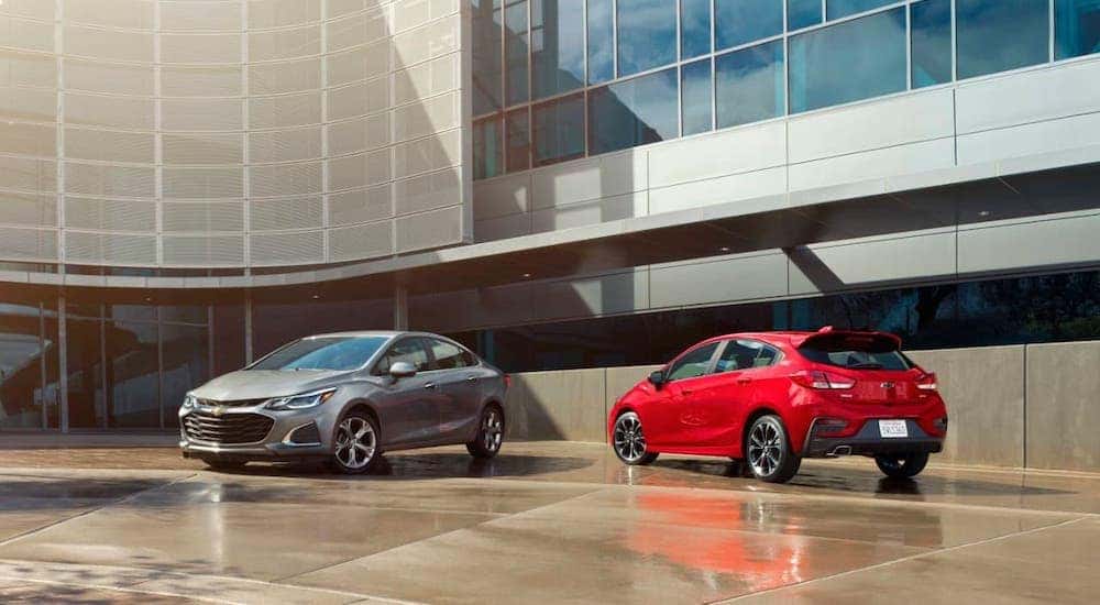 A gray 2019 Chevy Cruze sedan and a red hatchback are parked in front of a Louisville used car dealer.