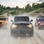 A yellow, a black , and a red 2021 Ford Bronco are shown driving down a dirt path in a 'V' formtion.