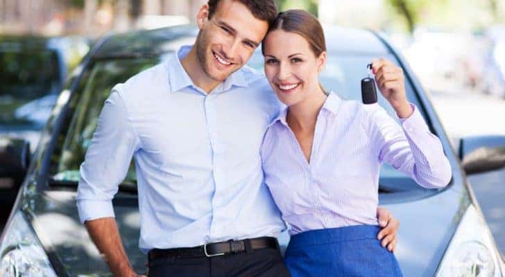 A smiling couple is holding up a car key while leaning on a grey car.