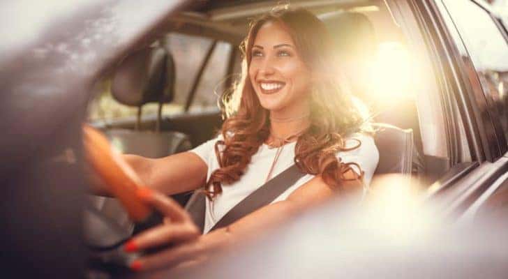 A woman is smiling while driving a vehicle, shown through the open driver's window.