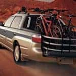 A tan and green 2005 Subaru Baja is driving dwon a dirt road with bikes in the back.