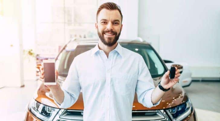 A man is in front of an orange SUV with a car key and his phone at a dealership.