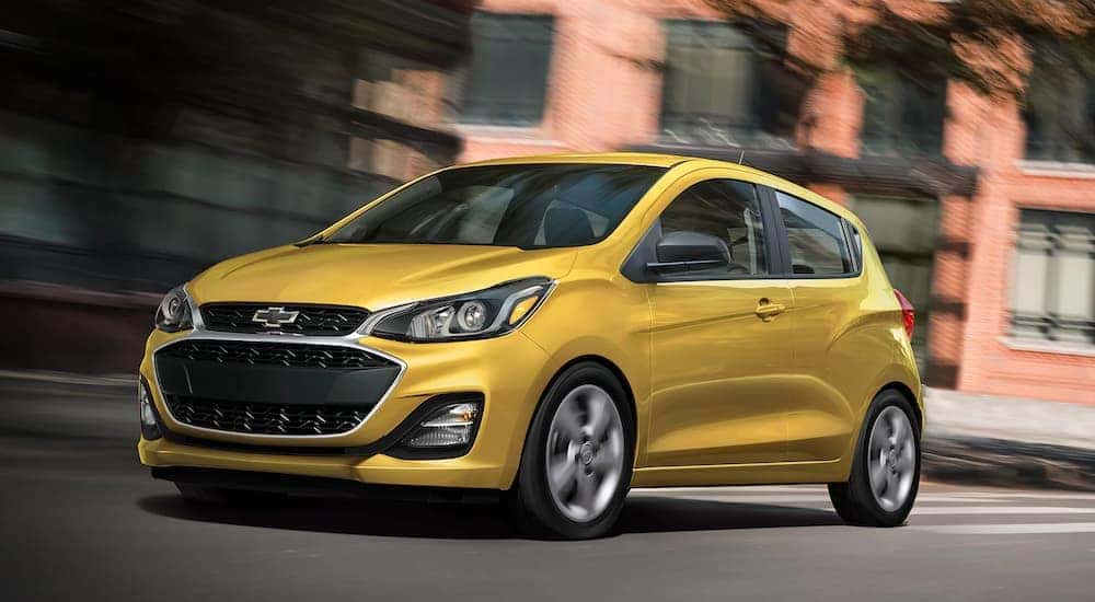 A yellow 2022 Chevy Spark is shown driving past a brick building.