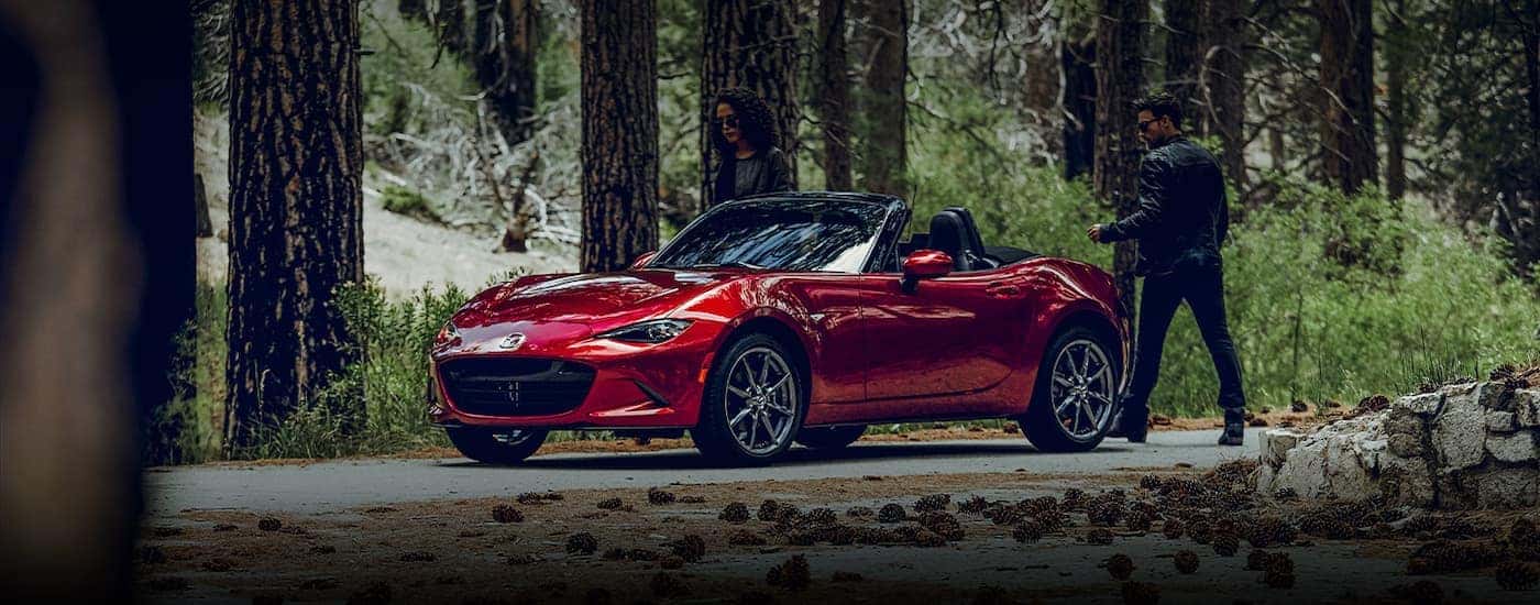 A red 2021 Mazda MX-5 Miata is shown from the side parked on a wooded road.