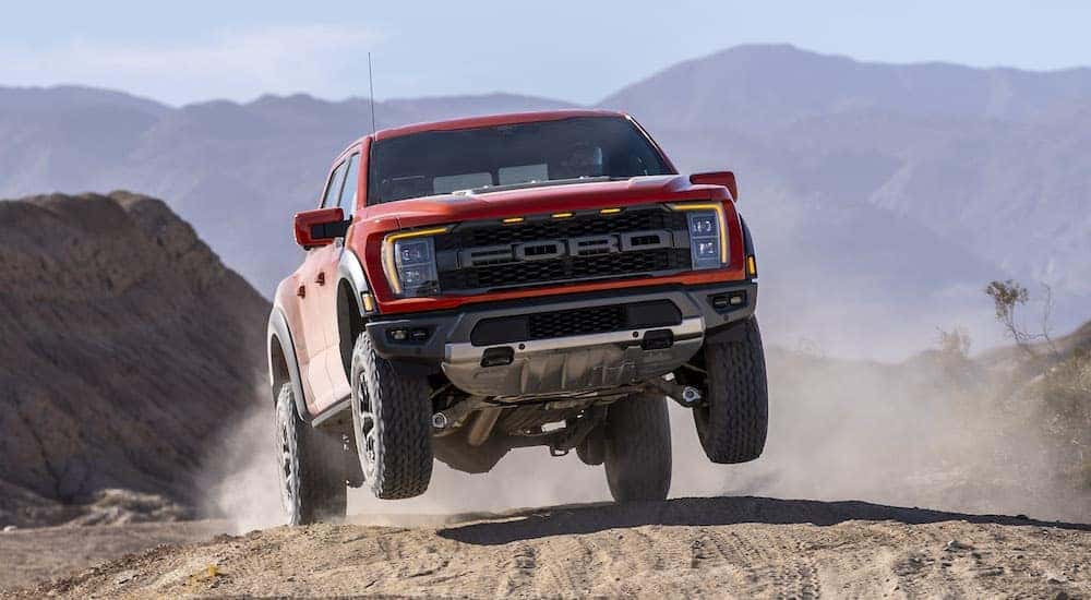 An orange 2021 Ford F-150 Raptor is shown from the front while catching air off of a sandy hill.