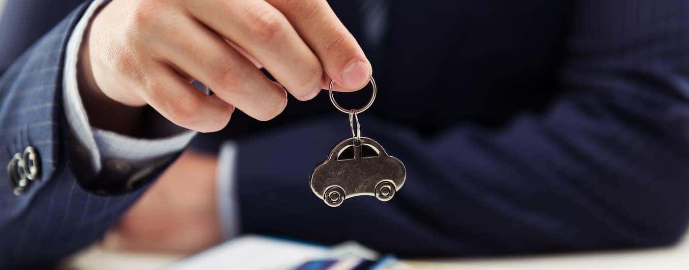 A close up of a person in a suit is shown holding a silver car-shaped keychain.