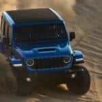 A blue 2024 Jeep Wrangler Rubicon is shown driving downhill on sand.