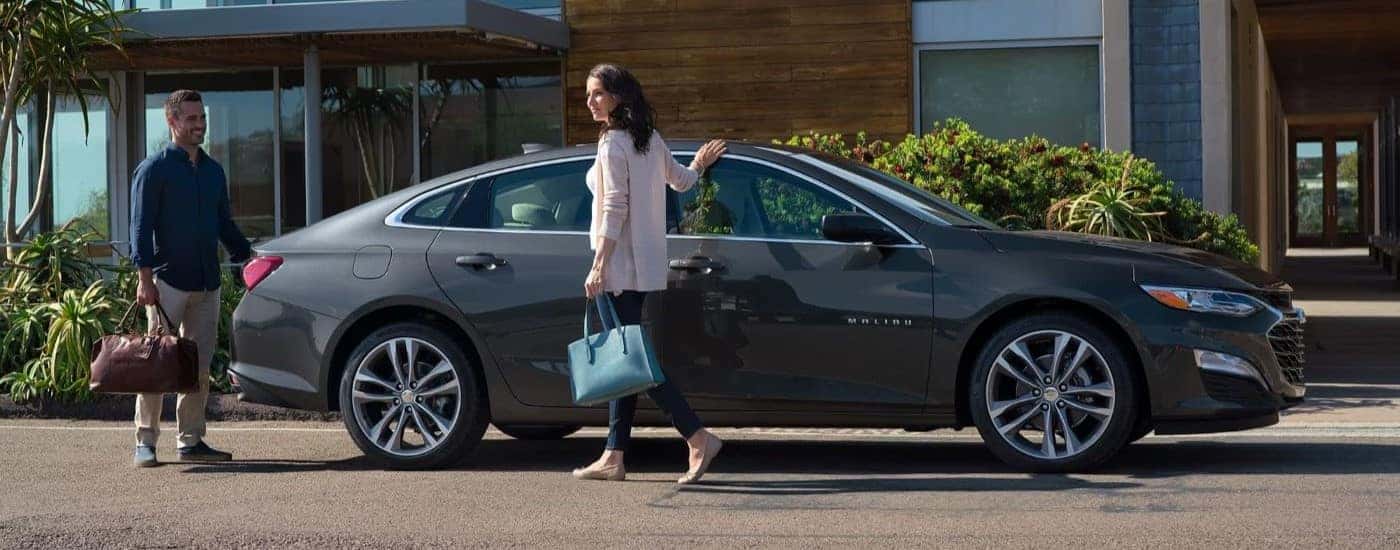A gray 2023 Chevy Malibu is shown parked near two people carrying bags.