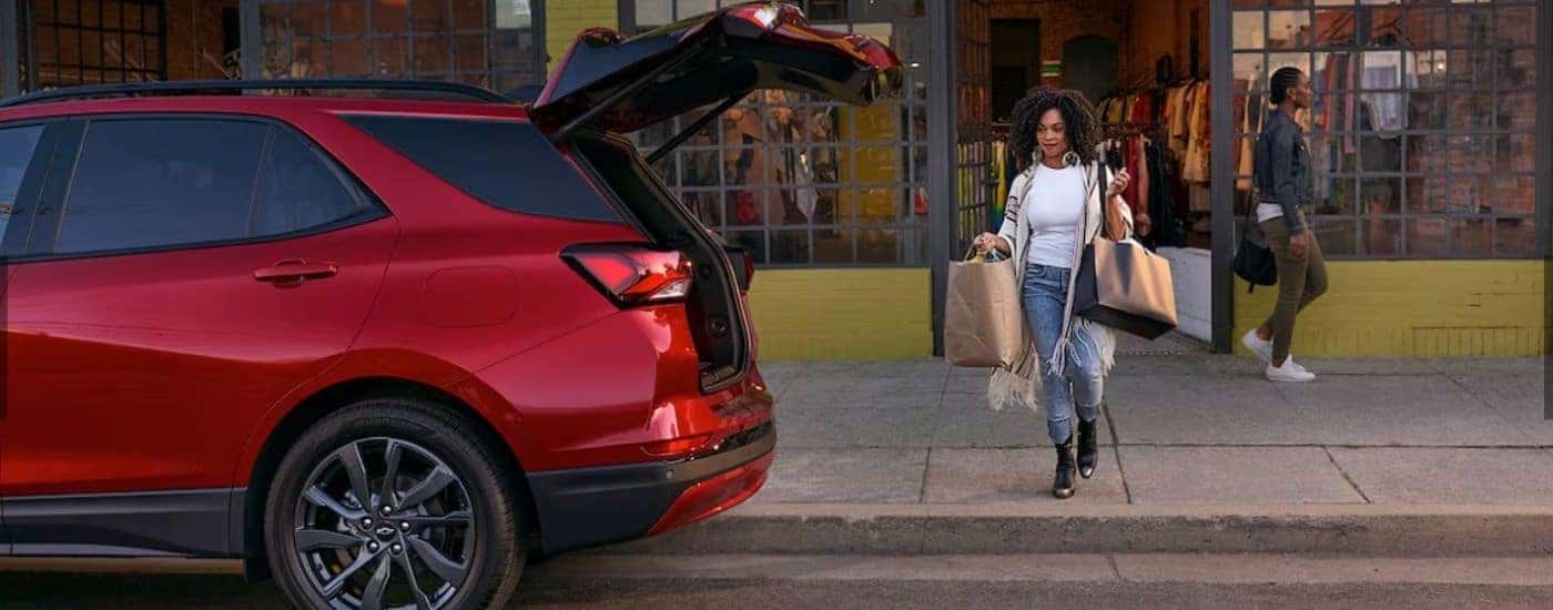 Side angle view of a woman carrying bags to the open hatchback of a red 2024 Chevy Equinox RS.