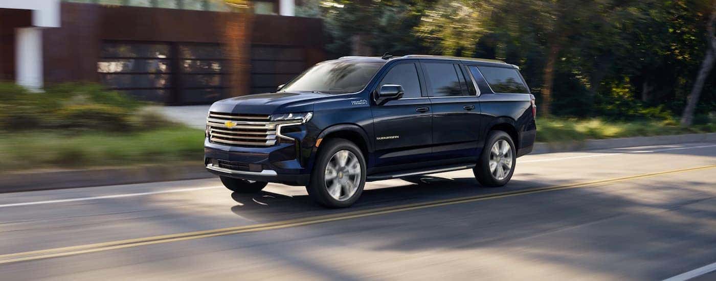 Left angled view of a blue 2021 Chevy Suburban High Country driving from a dealer with a Chevy Suburban for sale.