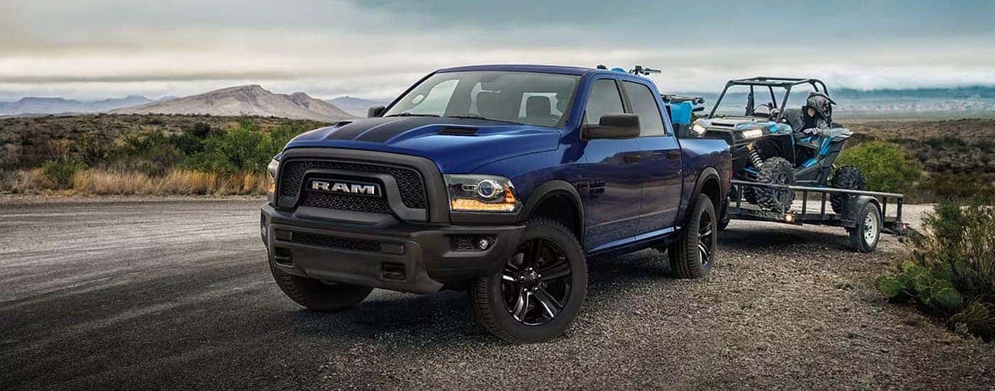 Left-angled view of a blue 2024 Ram 1500 towing an ATV from a dealer with a Ram 1500 for sale in Nashville.