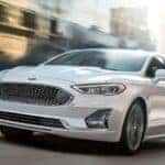 One of many popular CPO Fords for sale, a white 2020 Ford Fusion, is shown driving on a city street.