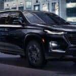 One of many popular CPO Chevys for sale, a black 2023 Chevy Traverse, is shown driving on a city street.