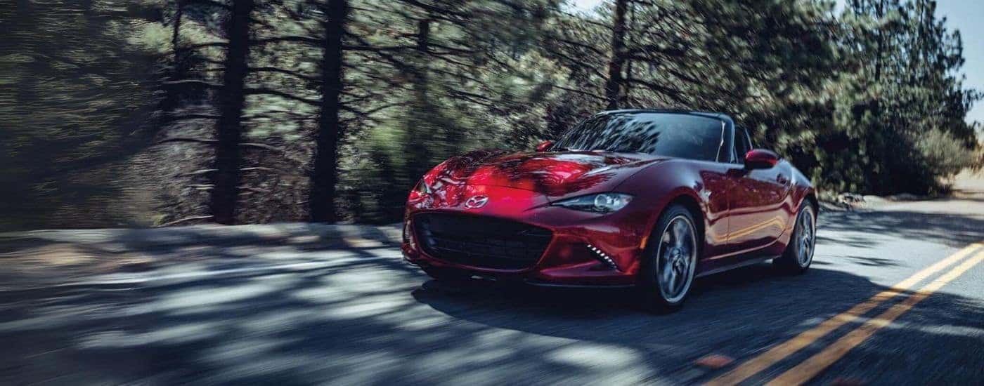 A red 2021 Mazda MX-5 Miata driving a tree-lined road.