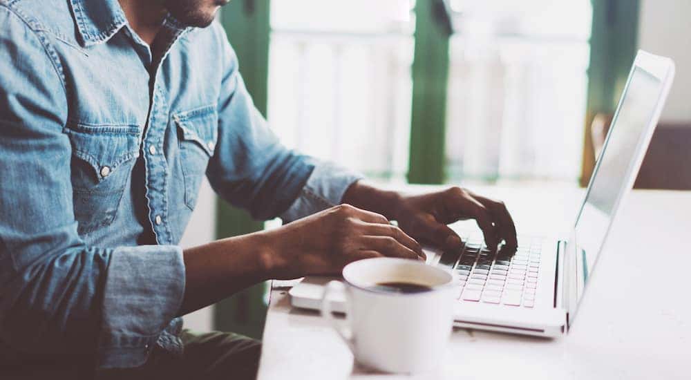 Man in a blue shirt sitting at a white table with a white cup of coffee, using a laptop
