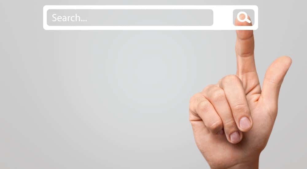 Hand up in front of a grey wall with the pointer finger touching a white "SEARCH" bar floating in the air