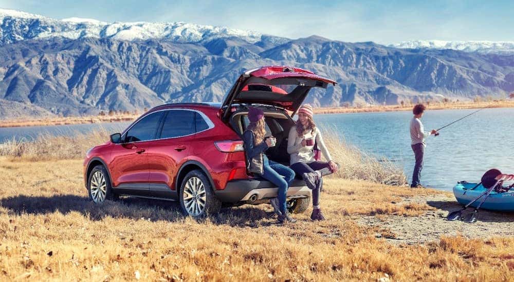 Red 2020 Ford Escape sitting next to a body of water and snowy mountains with the trunk open, two women sitting on the back, and a woman fishing in the background