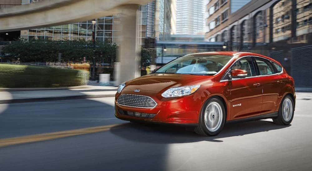 Red 2018 Ford Focus Electric driving under a bridge and next to tall corporate buildings