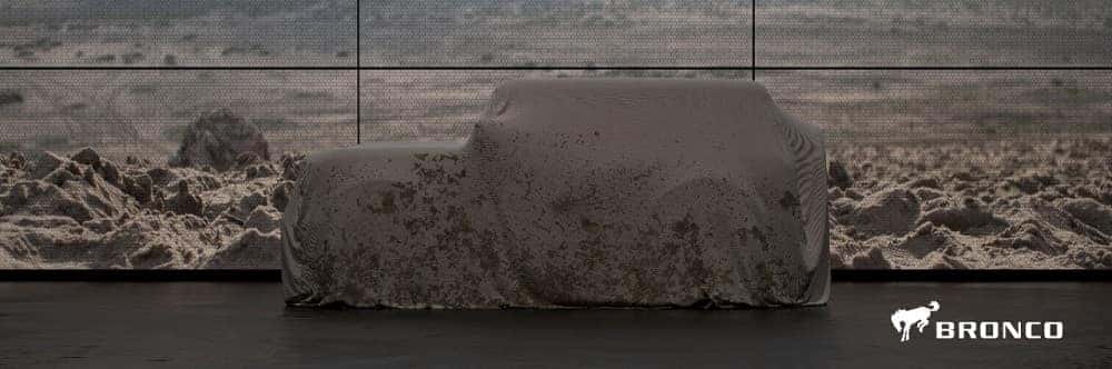 2020 Ford Bronco under a tan mud-splattered car cover with a wall of TVs in the background making a landscape picture of sand dunes 