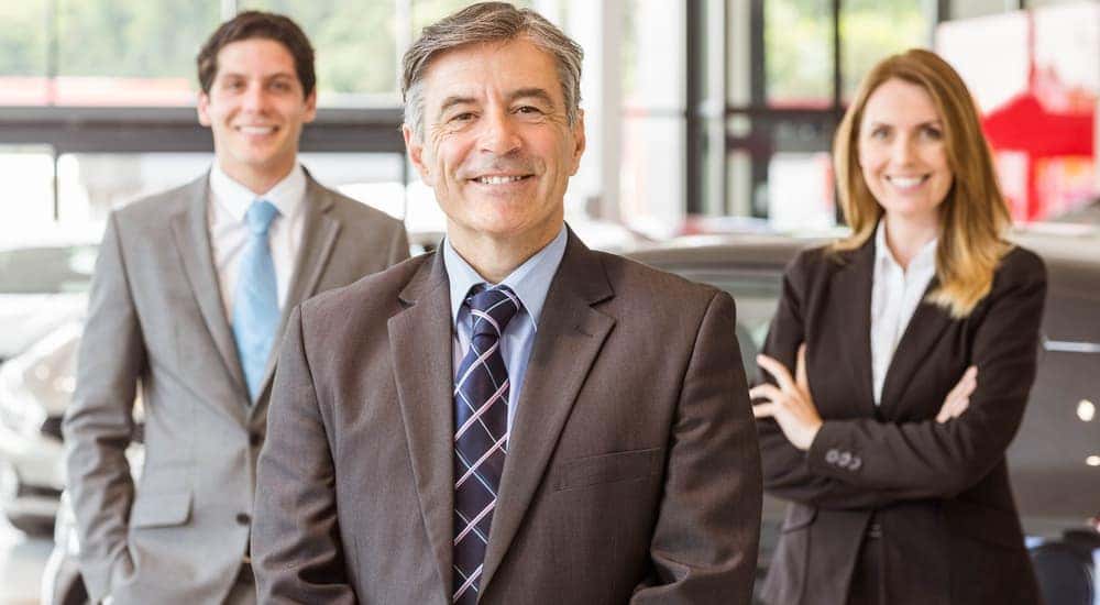 Three smiling sales people are standing in a local car dealers showroom in Albany, NY.