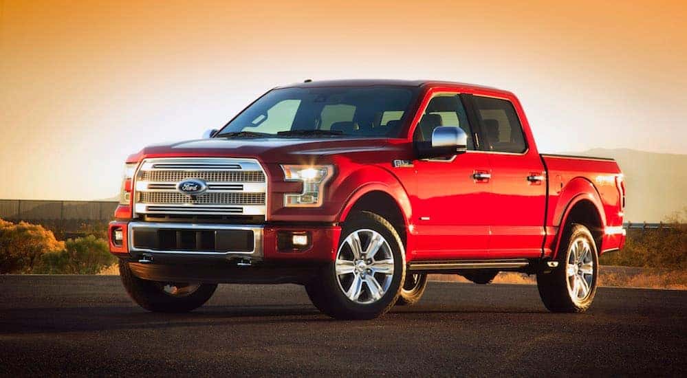 A red 2015 Ford F-150 is parked in an empty parking lot while the sun sets behind it.
