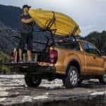 A man is taking his kayak off of the bedrails on his orange 2020 Ford Ranger next to a rocky river.
