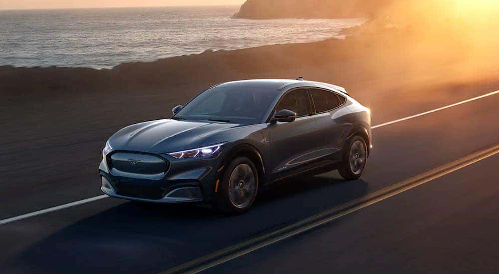 A grey 2021 Ford Mustang Mach-E is driving past the ocean at sunset.