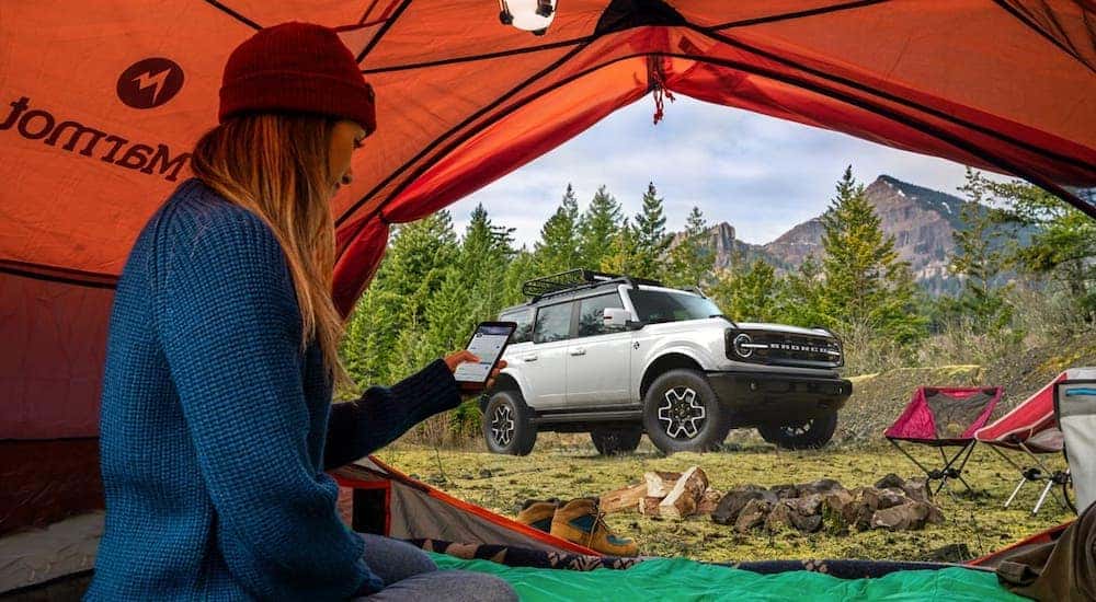 A woman is on her phone in a tent while a silver 2021 Ford Bronco Outer Banks 4-door from a Ford dealership near you is in the background.