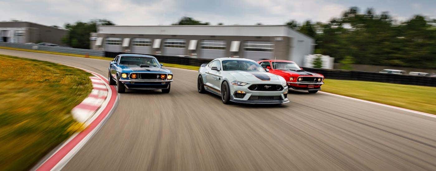 A white 2021 Ford Mustang Mach 1 is flanked by a blue and a red 1960s Mustang Mach 1s on a race track.