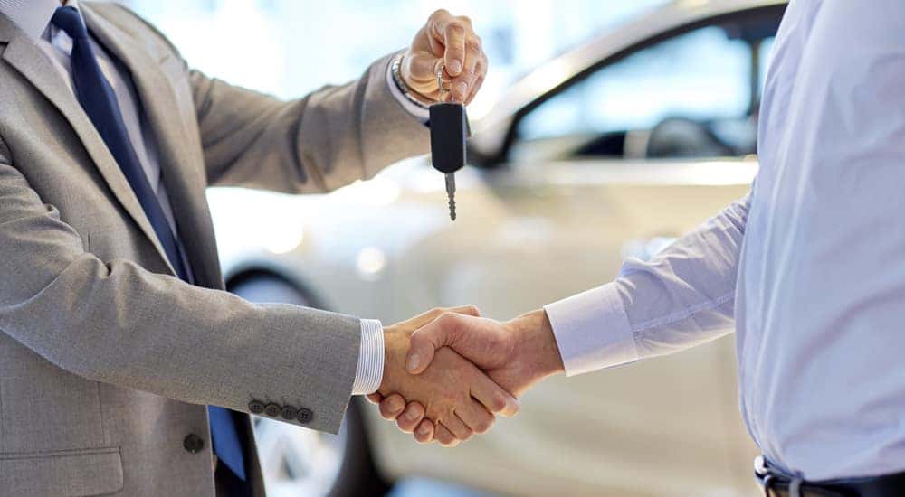 Man in a suit shaking hands with a man in a blue button-up shirt while handing him a car key in front of a white SUV