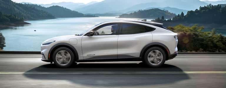 A white 2021 Ford Mustang Mach E is driving past a lake and mountains.