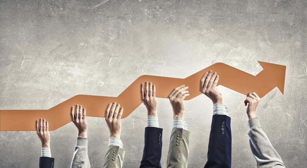 Hands from people in suits holding up an orange arrow that is moving to the right and upwards