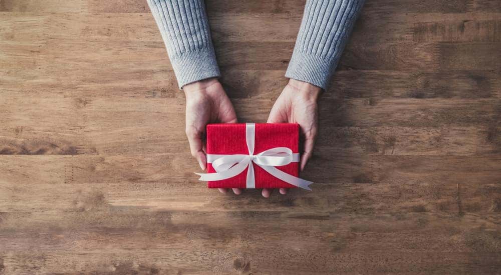 Hands holding a red box with a white ribbon around it on top of a brown wooden table