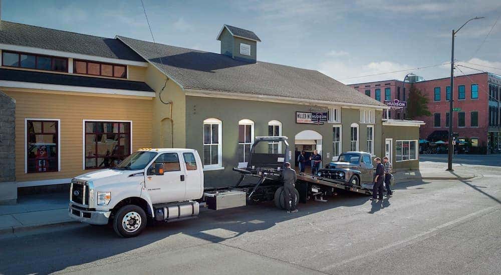 A white 2021 Ford F-650 is shown with its bed down to tow a classic black Ford pickup truck.