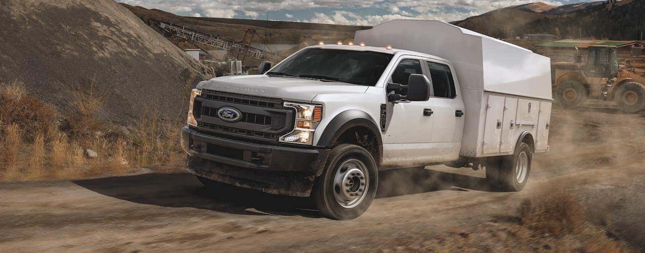 A white 2012 Ford F-450 with a utility upfit is driving on a dirt road through hills.