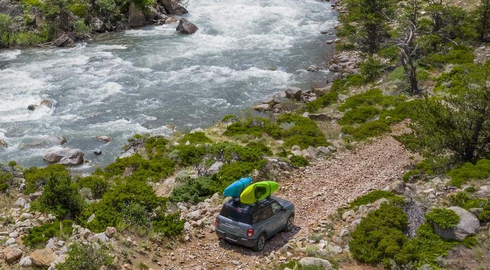 A blue 2021 Ford Bronco Sport is driving next to a river with two kayaks mounted on the roof.